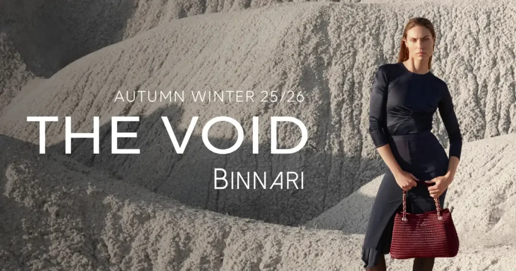 Mujer vestida con un elegante vestido negro, posando en un paisaje desértico con bolso rojo granate en mano, como parte de la campaña THE VOID de Binnari para bolsos otoño invierno 25/26.