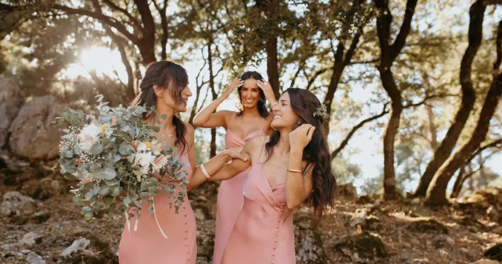 Tres damas de honor o invitadas con vestidos rosa satinado y ramo de flores en un entorno natural boscoso.